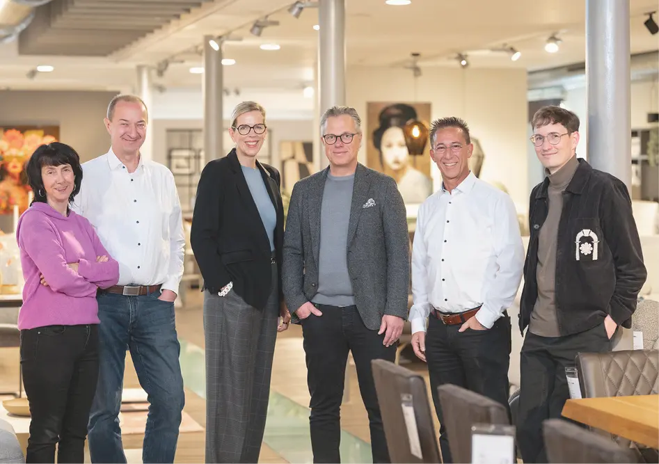 Sechs Personen stehen im Möbelhaus zwischen Esstisch und Stühlen, im Hintergrund Lampen und Wohnmöbel im Showroom